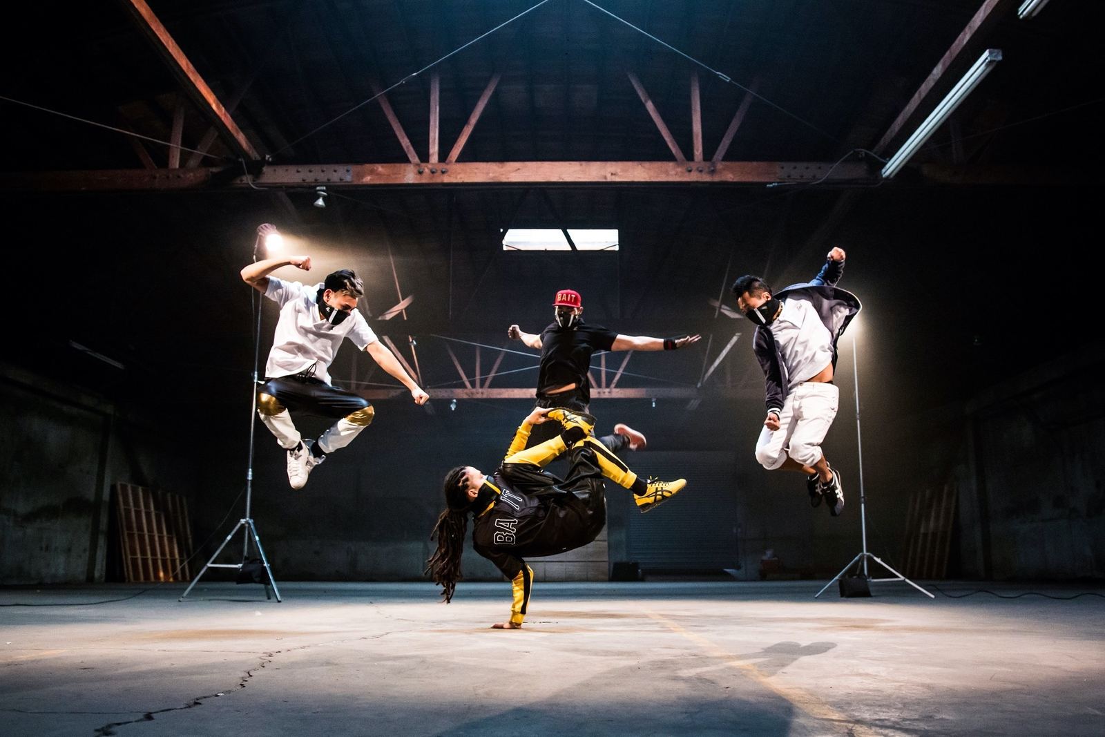 Four dancers in a warehouse: three in mid-air, one in yellow and black balancing on one hand