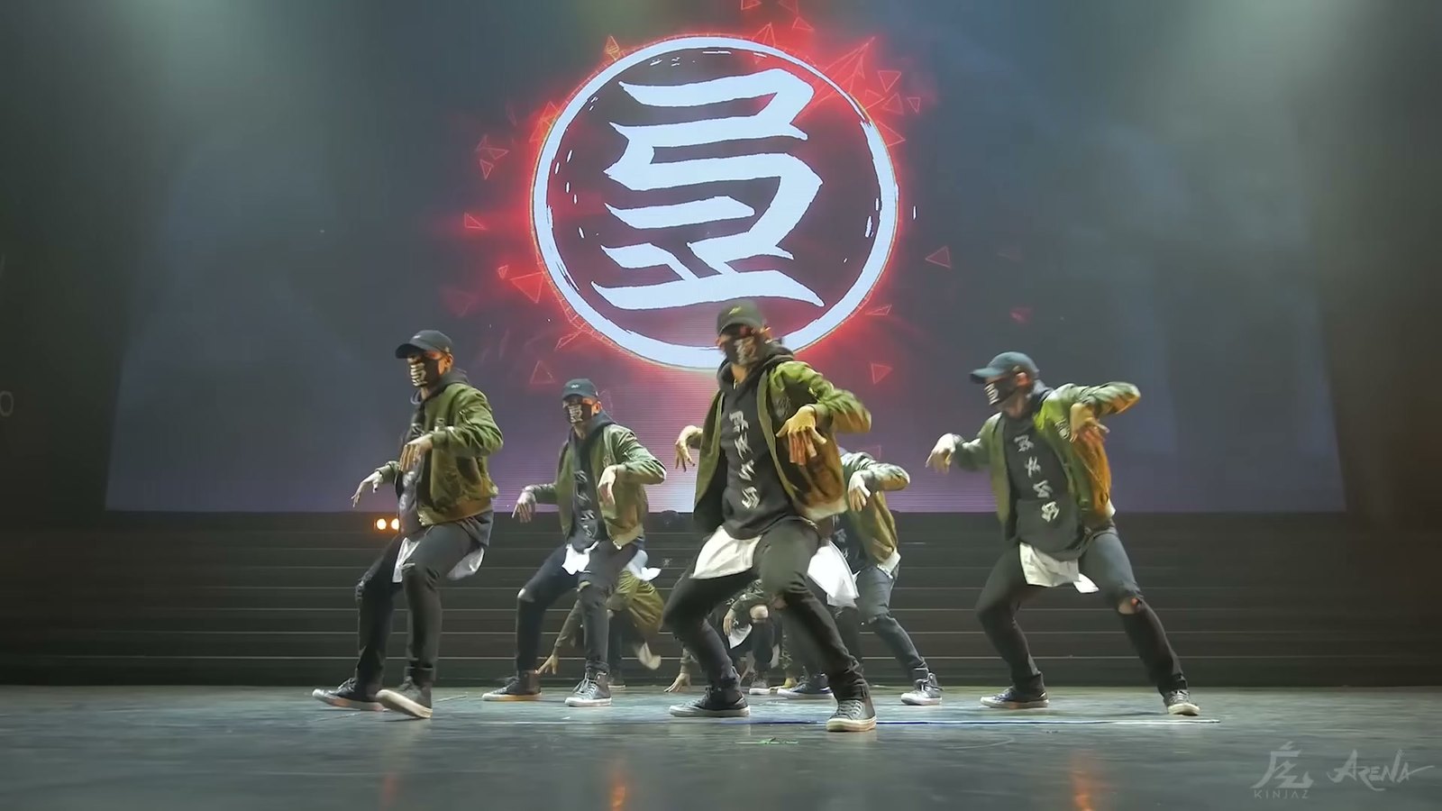 Masked Kinjaz dancers in olive jackets mid-move under a huge red Kinjaz mark on the Arena stage