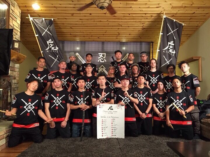 Twenty-some crew members in matching black shirts with red sashes in a cabin, between two Kinjaz banners, holding a signed welcome board
