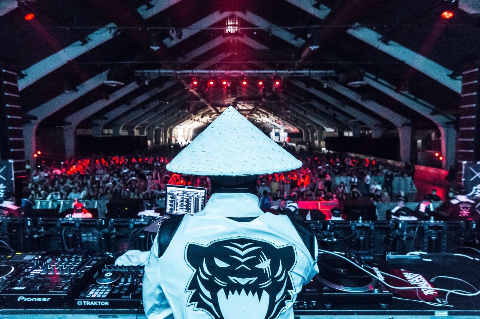 A figure in a rice hat and a Kinjaz jacket facing a festival crowd from the DJ booth