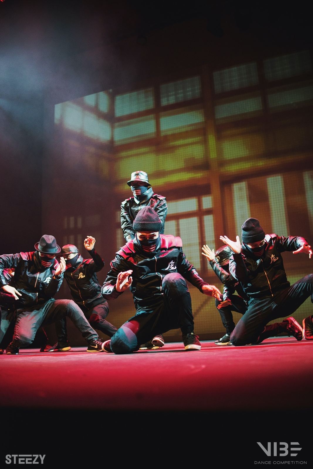 Masked dancers crouched in formation on a red-lit stage